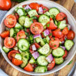 Cucumber tomato salad with sliced cucumbers, tomatoes, and fresh herbs in a white bowl on a wooden table