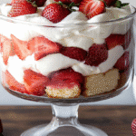 Layered trifle strawberry shortcake with pound cake, strawberries, pudding, and cream in a glass bowl on a wooden table.