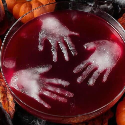 Spooky Halloween party punch in a clear bowl with fruit and gummy worms, styled with pumpkins and spider webs.